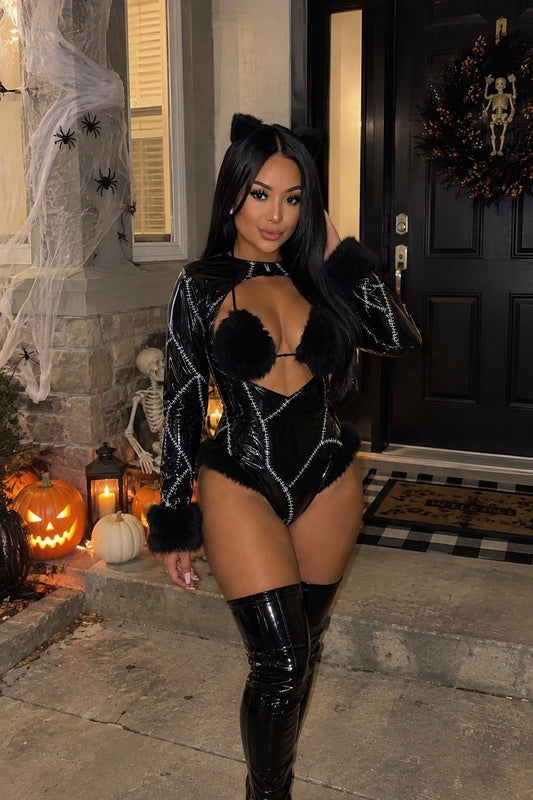 A woman posing in a black long-sleeve catgirl bodysuit with faux fur trim and silver rhinestone patterns, paired with black thigh-high boots, set against a Halloween decorated front porch.